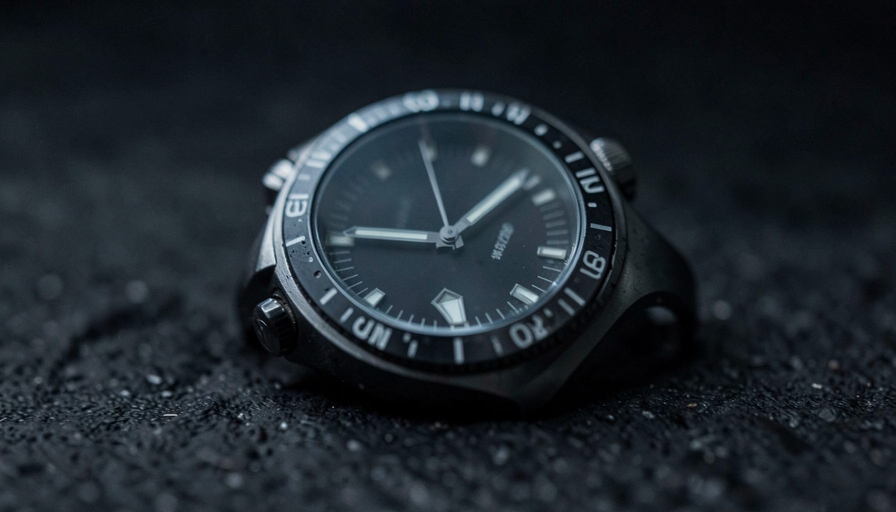 Close-up of a modern dive watch with a dark dial and unidirectional bezel, resting on a textured dark surface under moody lighting