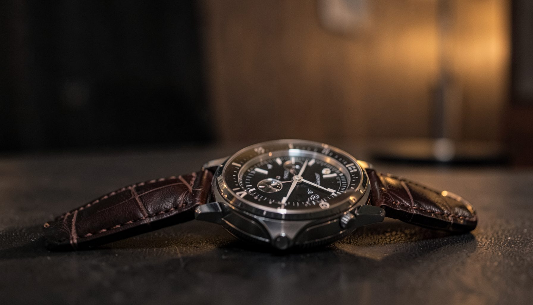 Softly lit interview setting with a luxury chronograph on a leather strap in the foreground, warm amber and charcoal tones