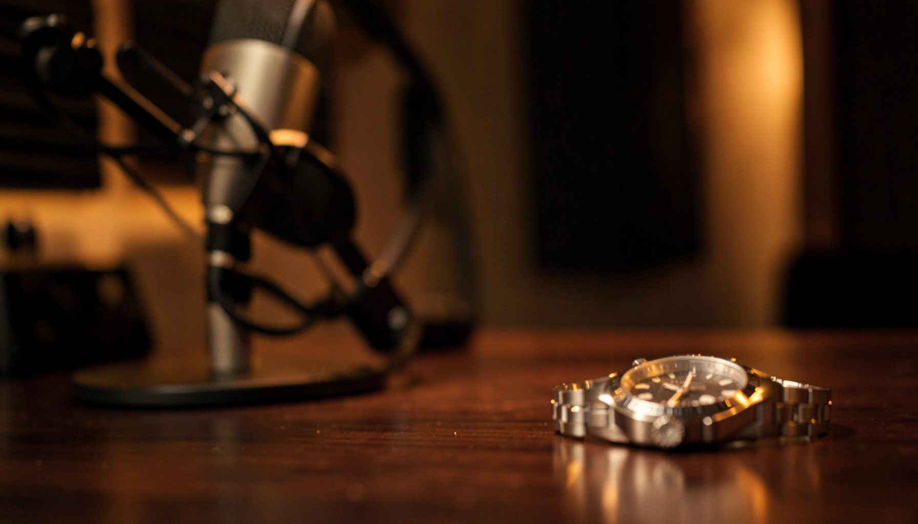 Warm-toned podcast studio with a professional microphone on a desk, soft amber lighting, and a blurred luxury watch resting nearby